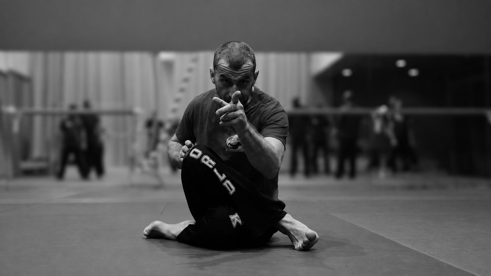 Pratiquant de penchak silat assis au sol, la main tendue vers l'objectif, en noir et blanc